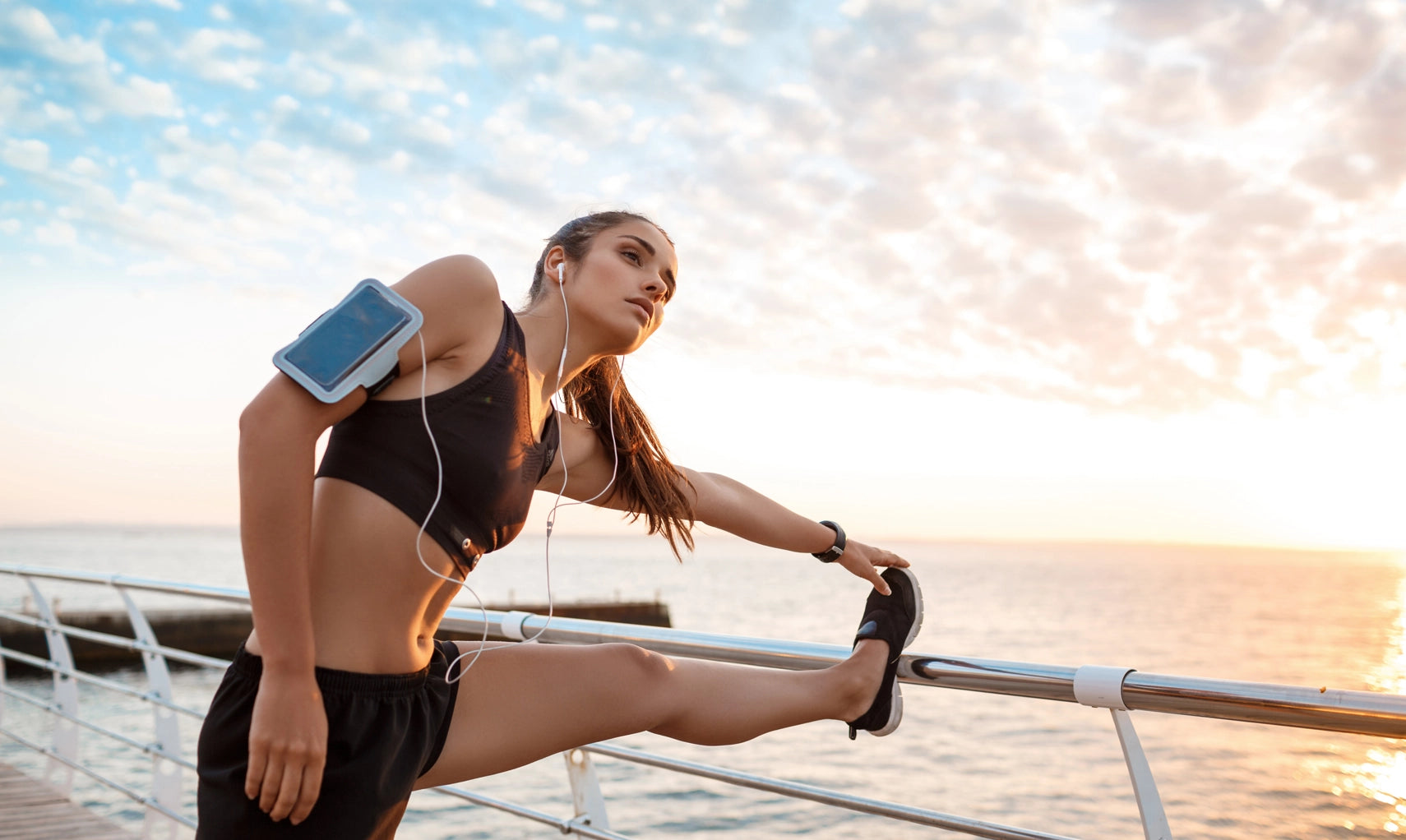 Woman stretching outdoors with a smartphone in an arm band, sunset in the background