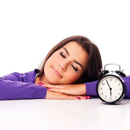Woman resting her head on a table with an alarm clock next to her, symbolizing the passage of time or the need for sleep.