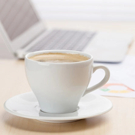 White coffee cup with a saucer on a desk with a blurred laptop in the background
